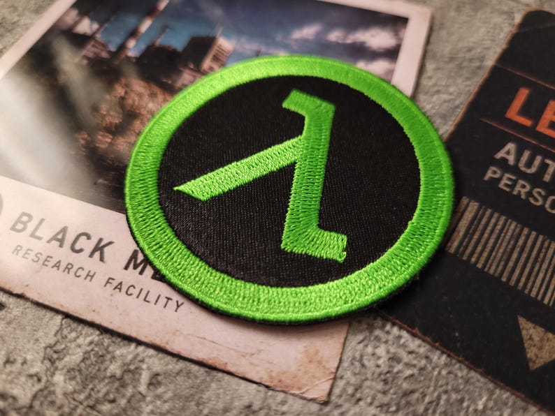 May include: A circular embroidered patch featuring a bright neon green lambda symbol centred on a solid black background, surrounded by a thick neon green stitched border. The patch is circular in shape with a textured thread finish throughout.