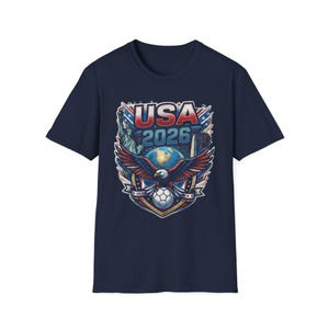 May include: A navy blue short-sleeve t-shirt featuring a graphic print of a bald eagle with wings spread, a soccer ball, the Statue of Liberty, the Washington Monument, a globe, and the text USA 2026 in red, white, and blue patriotic colors.