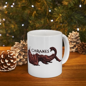 Könnte beinhalten: Ein weißer Keramikbecher mit einer dunkelroten Illustration eines Drachen mit großen Flügeln und langem Hals. Der Schriftzug "CARAXES" ist in einer dunkelroten Serifenschrift über dem Rücken des Drachen aufgedruckt. Der Becher hat einen Henkel.