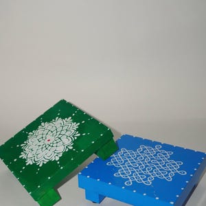 Peut inclure: Deux tabourets bas carrés en bois avec des pieds courts. Un tabouret est peint en vert avec un motif mandala blanc et un petit point rouge au centre. L'autre tabouret est peint en bleu avec un motif kolam géométrique blanc et des points blancs.