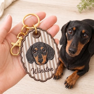 Puede incluir: Un llavero bordado de forma ovalada con el retrato de un perro salchicha negro y fuego y el nombre Winston en letra cursiva. El fondo tiene rayas verticales beige y blancas con un borde festoneado marrón y una anilla de metal dorado.
