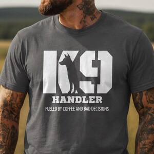 Puede incluir: Una camiseta gris oscuro de manga corta con un gráfico blanco que incluye el texto K9 HANDLER y FUELED BY COFFEE AND BAD DECISIONS, con la silueta de un perro integrada en la letra K, junto con una gorra de béisbol negra.