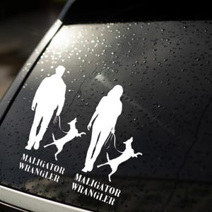 May include: A white vinyl decal applied to a dark car window, featuring two silhouettes of people walking dogs on leashes. Below each figure, the text reads MALIGATOR WRANGLER in a bold, serif font. The design depicts two active dogs jumping.
