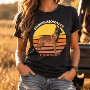 Puede incluir: Una camiseta negra de manga corta con el gráfico de un perro pastor belga malinois marrón frente a un círculo de atardecer amarillo y naranja. El texto "PROFESSIONALLY" está arqueado sobre el perro y "UNHINGED" impreso debajo.