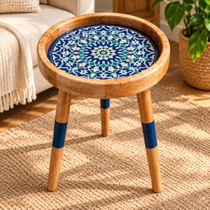 May include: A round wooden side table with three legs. The tabletop features a circular inlay with a detailed blue, green, and white geometric mandala pattern. The legs are light brown wood with a thick horizontal band of dark blue paint.