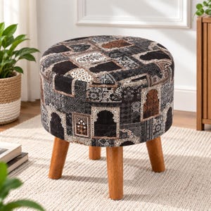 May include: A round ottoman featuring a dark, multi-patterned fabric upholstery with intricate geometric and architectural motifs in shades of black, brown, and grey. The piece is supported by four sturdy, light-toned wooden legs.