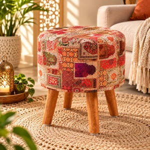 Puede incluir: Un taburete redondo con una vibrante tapicería de tela de retazos con intrincados patrones geométricos y florales en tonos rojo, naranja y morado. El asiento se apoya sobre cuatro patas de madera clara y robusta que se estrechan hacia abajo.