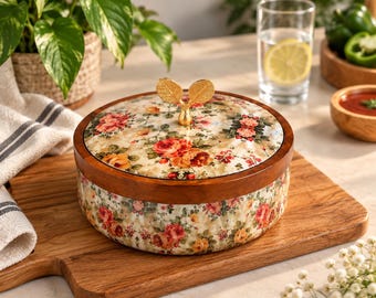 Puede incluir: Un recipiente redondo de cerámica con un estampado floral de rosas rojas, naranjas y amarillas sobre fondo crema. Tiene un borde de madera marrón oscuro y una tapa de cerámica a juego rematada con un asa de metal dorado con forma de dos hojas.