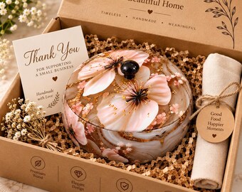 Puede incluir: Un recipiente redondo de resina con detalles florales rosas y láminas de oro, con una tapa de pomo negro. El artículo está en una caja de regalo de cartón con una tarjeta de agradecimiento, una servilleta de tela beige enrollada y flores secas.