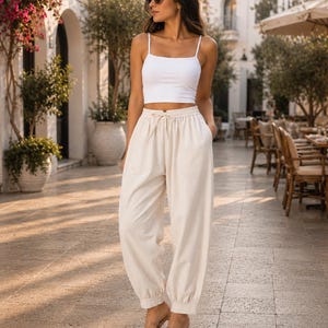 Può includere: Un top corto bianco senza maniche con spalline sottili abbinato a pantaloni da jogging color crema a vita alta con elastico in vita, coulisse e polsini arricciati alle caviglie, indossato con sandali con tacco trasparenti.