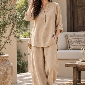 Op de afbeelding: Een beige tweedelige set bestaande uit een tuniek met lange mouwen, een opstaande kraag, een gedeeltelijke knoopsluiting en opgerolde mouwen, gecombineerd met een bijpassende wijde broek van een gestructureerde, gekreukte stof.