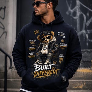 May include: A black hooded sweatshirt featuring a graphic of a teddy bear wearing a leather jacket, cap, and sunglasses. The print includes the text "SELF MADE," "BUILT DIFFERENT," and motivational phrases like "DISCIPLINE TODAY" and "NO RISK NO STORY."