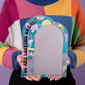 May include: A colorful, abstract arch-shaped mirror with the text "YOU ARE LOOKING GREAT!" printed on the side. The mirror has a light blue background with pink, yellow, and white abstract shapes.