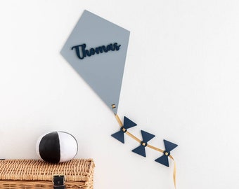 May include: A light blue kite with the name "Thomas" written on it. The kite is hanging on a white wall with a gold string attached to it. There are three blue bow-shaped decorations on the string.