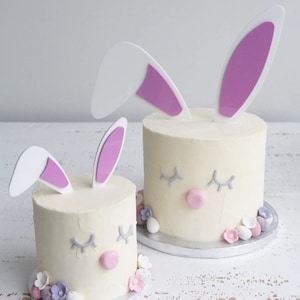 Puede incluir: Dos pasteles blancos decorados con orejas de conejo rosas, pestañas grises y narices rosas. Los pasteles están rodeados de flores moradas y blancas y huevos de caramelo blancos.