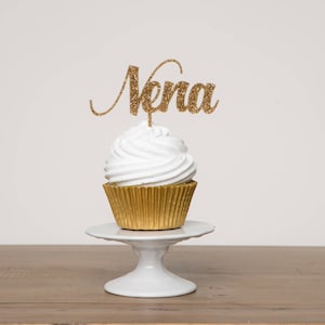 Pode incluir: Um cupcake branco com cobertura dourada com glitter coroado com um topper de cupcake dourado com glitter que diz "Nena" em um suporte de bolo branco.