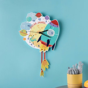 Op de afbeelding: Een kleurrijke raketvormige klok met een maan gezicht en planeten. De wijzerplaat van de klok heeft de tekst "Oscar James" en heeft een rood, wit en blauw kleurenschema. De klok hangt aan een lichtblauwe muur.