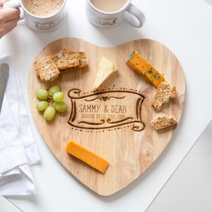 Può includere: Un tagliere per formaggi in legno a forma di cuore con l'iscrizione "Sammy & Dean Sharing Cheese Since 2019". Il tagliere è pieno di formaggio, cracker e uva.