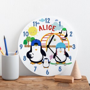 Puede incluir: Un reloj redondo blanco con números azules y un diseño colorido con tres pingüinos, un sol, un árbol verde y un parque infantil rojo y amarillo. El reloj dice "ALICE" en letras rojas.