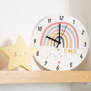Op de afbeelding: Een witte ronde klok met een regenboogontwerp en zwarte wijzers. De klok heeft een witte achtergrond met een regenboogontwerp en zwarte wijzers. Het wijzerplaat van de klok heeft de cijfers 1 tot en met 12 en het woord "ELODIE" erop geschreven. De klok staat op een houten plank met een geel stervormig nachtlampje.
