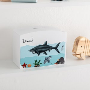 May include: White wooden piggy bank with a blue and white ocean scene featuring a shark, starfish, and seaweed. The name "Daniel" is written on the front of the bank.
