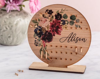 May include: A round wooden earring holder with a floral design and the name "Alison" printed on it. The holder has multiple holes for earrings and is on a small wooden stand.
