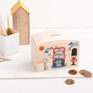 Könnte beinhalten: Hölzernes Sparschwein mit einem roten Doppeldeckerbus und einem britischen Wachmann auf der Vorderseite. Der Bus hat den Schriftzug "Murky" an der Seite. Vor dem Sparschwein liegen Münzen auf dem Tisch.