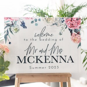 May include: A white wedding welcome sign with a floral border in shades of pink, blue, and green. The sign reads "Welcome to the wedding of Mr. and Mrs. MCKENNA Summer 2023".