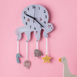 May include: A light grey octopus-shaped clock with a white face and black hands. The clock has the name "Sienna" written on it. The clock has five dangling charms: a seahorse, a mushroom, a plant, a star, and a goose.
