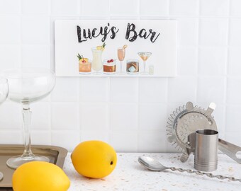 May include: A white sign with the text "Lucy's Bar" in black lettering. The sign features a row of six watercolour illustrations of different cocktails.