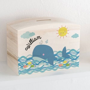 Puede incluir: Una alcancía de madera con un diseño de ballena y peces. La alcancía está pintada de azul, amarillo y blanco. El nombre "william" está escrito en el lateral de la alcancía.