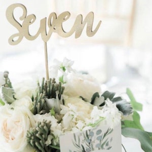 以下が含まれることがあります： 「Seven」と書いた、金の色の木製のテーブルナンバー。花束の中に置かれています。花束は白い花と緑の葉でできています。