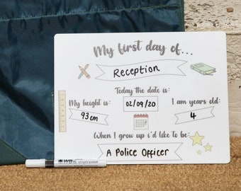 May include: A white dry-erase board with the text "My first day of... Reception" and other details, including height (93 cm) and age (4). A dark green satchel bag is behind the board, and a black marker is in front.
