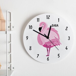 May include: A white round clock with black hands and black numbers. The clock features a pink flamingo illustration and the name "SARA" printed on the face.