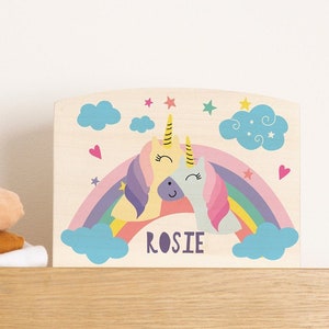 May include: Wooden sign with a rainbow and two unicorns hugging. The sign has the name "ROSIE" written on it.