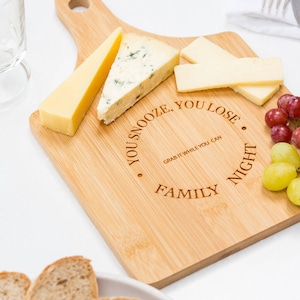 Può includere: Un tagliere di legno per formaggi con manico, con la scritta "You snooze, you lose. Grab it while you can. Family night." Il tagliere è pieno di formaggio, uva e pane.