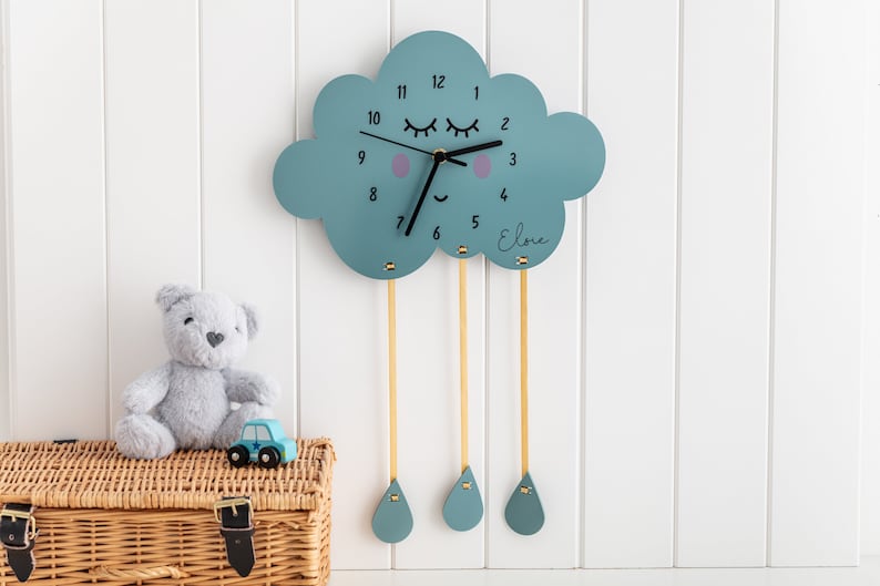 May include: A blue wooden cloud-shaped clock with a smiling face and eyelashes. The clock has gold hands and black numbers. The name "Elodie" is written on the clock. Three gold strings hang from the bottom of the cloud, each with a blue raindrop at the end.