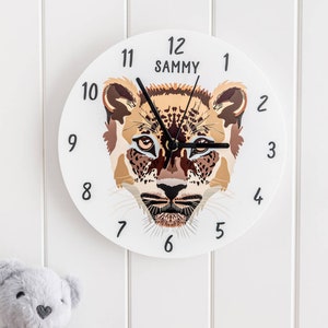 Puede incluir: Reloj redondo blanco con una cara de guepardo en el centro. La esfera del reloj tiene números negros del 1 al 12 y el nombre "SAMMY" en la parte superior.