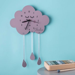 May include: A pink cloud-shaped clock with a smiling face and eyelashes, with the name "Charlie" written below the clock face. Three pink raindrops hang from the cloud, each attached by a white string.