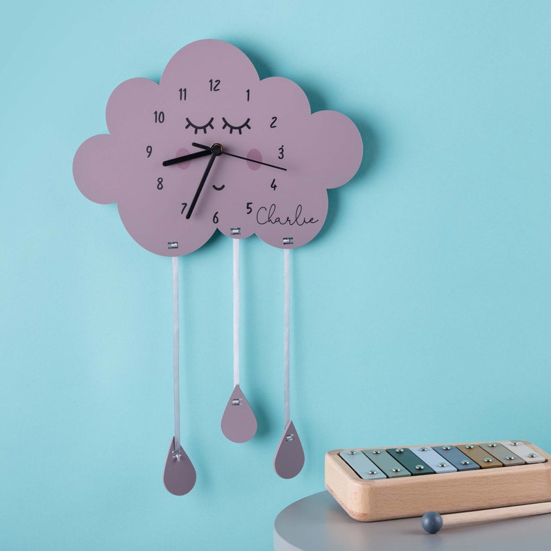 May include: A pink cloud-shaped clock with a smiling face and eyelashes, with the name "Charlie" written below the clock face. Three pink raindrops hang from the cloud, each attached by a white string.