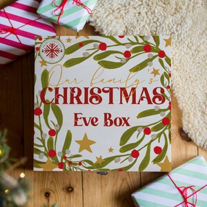 Peut inclure: Une boîte en bois blanche avec un design de boîte de Noël du réveillon. La boîte a un motif de gui rouge et vert avec des étoiles dorées. Le texte sur la boîte indique "Notre boîte de Noël du réveillon de famille".