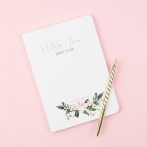 Könnte beinhalten: Ein weißes Notizbuch mit dem Text "Natalie Jones Bride to Be" und dem Datum "15.06.2019" auf der Vorderseite. Das Notizbuch hat ein florales Design mit rosa Blumen und grünen Blättern. Ein goldener Stift liegt auf dem Notizbuch.