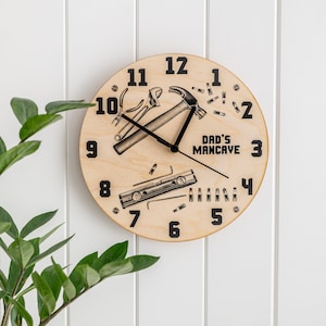 Puede incluir: Un reloj redondo de madera con números y manecillas negros. La esfera del reloj presenta un martillo, una llave inglesa, un nivel y clavos. El texto "Dad's Mancave" está impreso en la esfera del reloj.