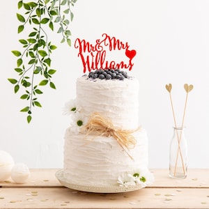 May include: A two-tiered white wedding cake with blueberries and a red acrylic cake topper that says "Mr. & Mrs. Williams" with a heart.
