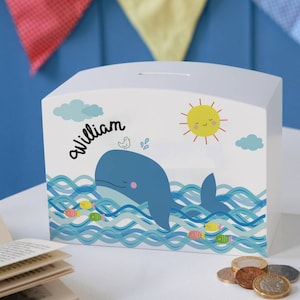 May include: A white, rectangular money box with a whale illustration. The box features a blue whale in blue ocean waves, with a smiling sun and the name "William" in black script. Colorful fish and clouds decorate the box.