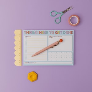 Pode incluir: Um bloco de notas azul e branco com o texto "Things I Need To Get Done" em letras arco-íris. O bloco de notas tem uma lista de verificação, uma seção de notas de procrastinação e uma seção de contato. Uma caneta rosa com uma tampa laranja está no bloco de notas. Há um objeto hexagonal amarelo, um rolo de fita adesiva e um par de tesouras turquesa sobre um fundo roxo.