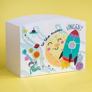 May include: White wooden piggy bank with a space-themed design. The design features a rocket, a moon, stars, planets, and an astronaut holding balloons. The text "to the moon" and the name "VINCENT" are printed on the bank.