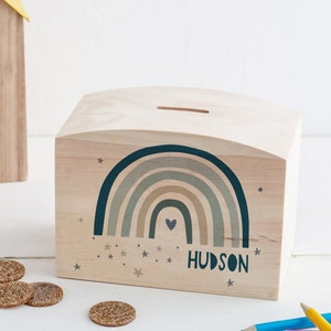 Könnte beinhalten: Hölzerne Regenbogen-Sparbüchse mit einem Schlitz oben. Der Regenbogen ist in Grüntönen und Braun mit einem kleinen Herzen in der Mitte. Die Worte "HUDSON" sind auf der Vorderseite der Sparbüchse aufgedruckt.