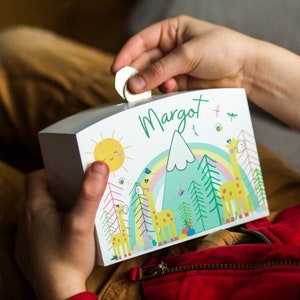 May include: A white wooden money box with a curved top. The box is decorated with a colorful illustration of giraffes, a rainbow, a mountain, and a smiling sun. The name "Margot" is written in green script on the front. A coin is being inserted into the slot.