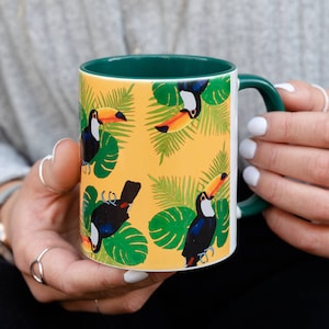 Könnte beinhalten: Eine gelbe Keramiktasse mit grünem Rand und Henkel zeigt ein sich wiederholendes Muster aus Tukanen und tropischen Blättern. Die Tukane sind schwarz, weiß und orange mit gelben Schnäbeln.